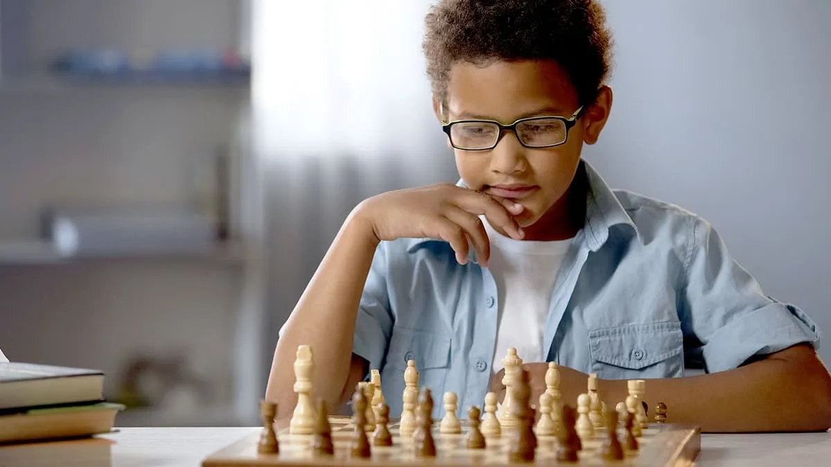 Boy wearing glasses in a blue shirt concentrating on a chess game at a table indoors.