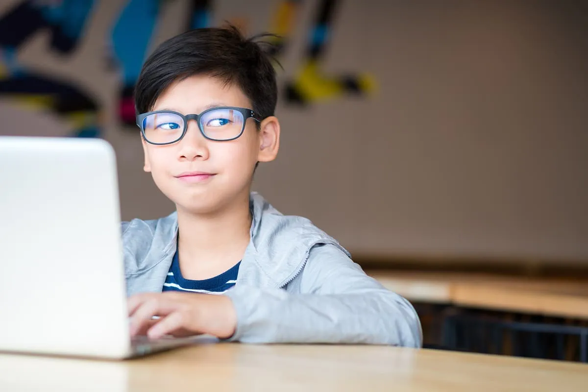 Young boy wearing glasses and a jacket, using a laptop while looking thoughtfully to the side.