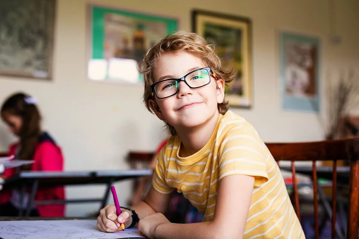 Smiling child with glasses and curly hair in a yellow striped shirt writing with a purple pencil in class.
