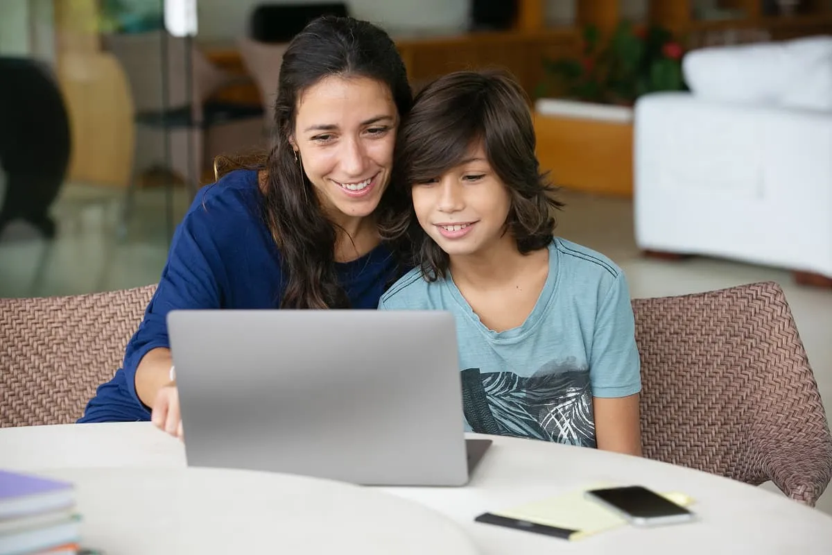 Woman and boy sitting together smiling while looking at a laptop screen on a white table.