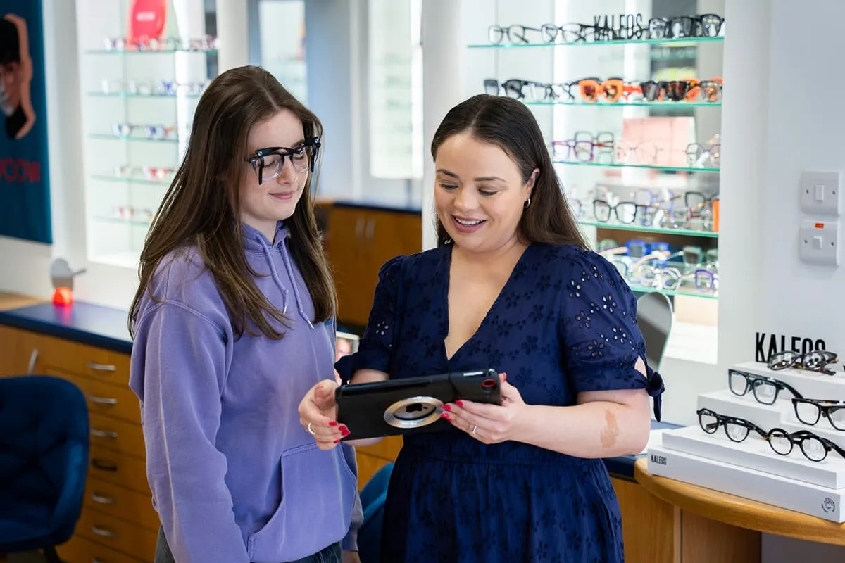 Smiling optician showing a tablet to a young woman wearing test glasses inside an eyewear store.