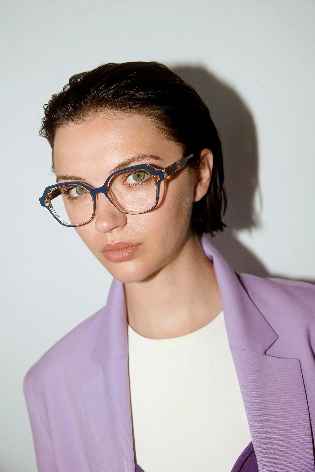 Woman with short dark hair wearing designer glasses with a blue frame and a lavender blazer.