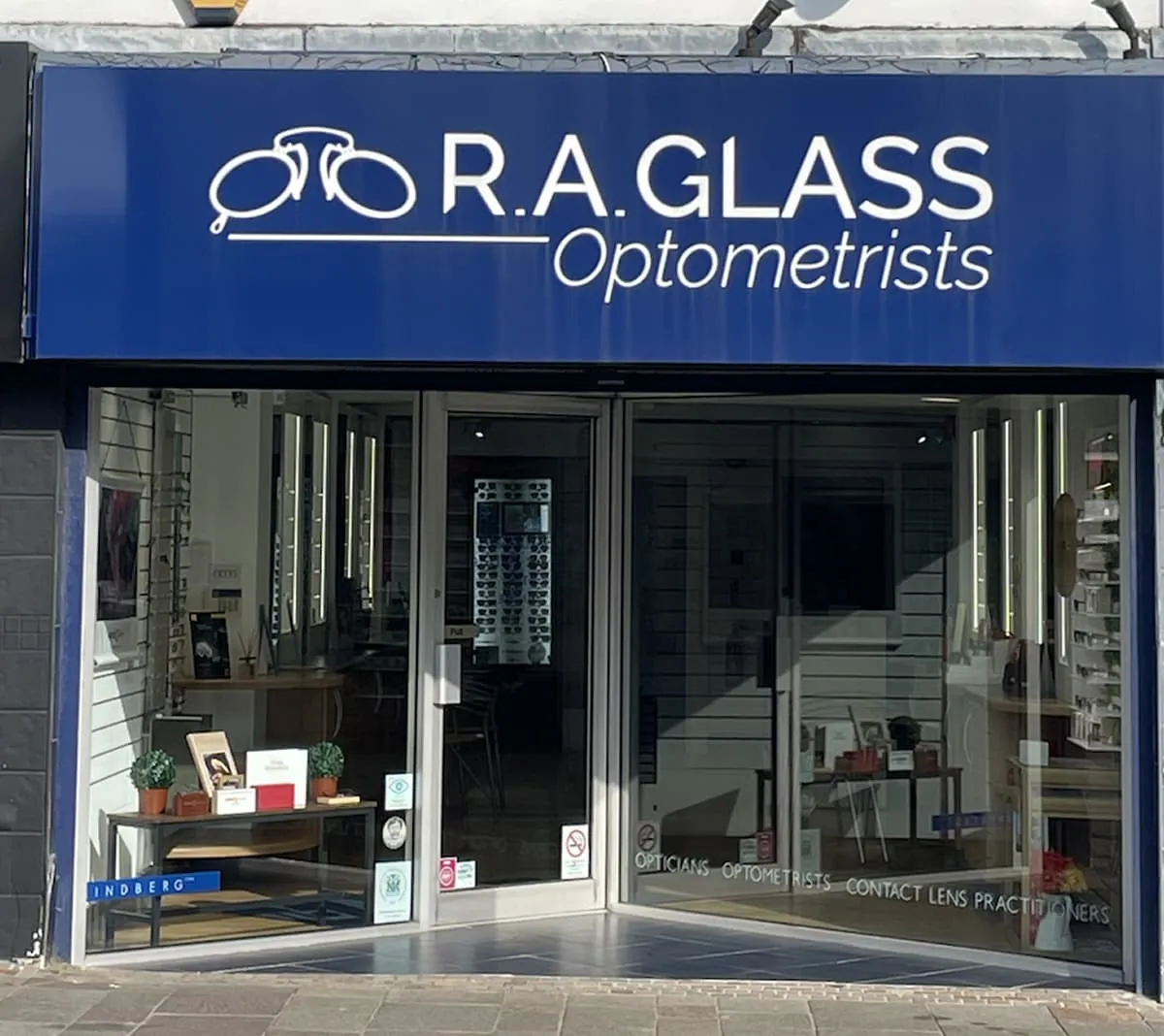 Storefront of R.A. Glass Optometrists with large blue sign featuring eyeglasses icon and glass doors showing eyeglass displays inside.