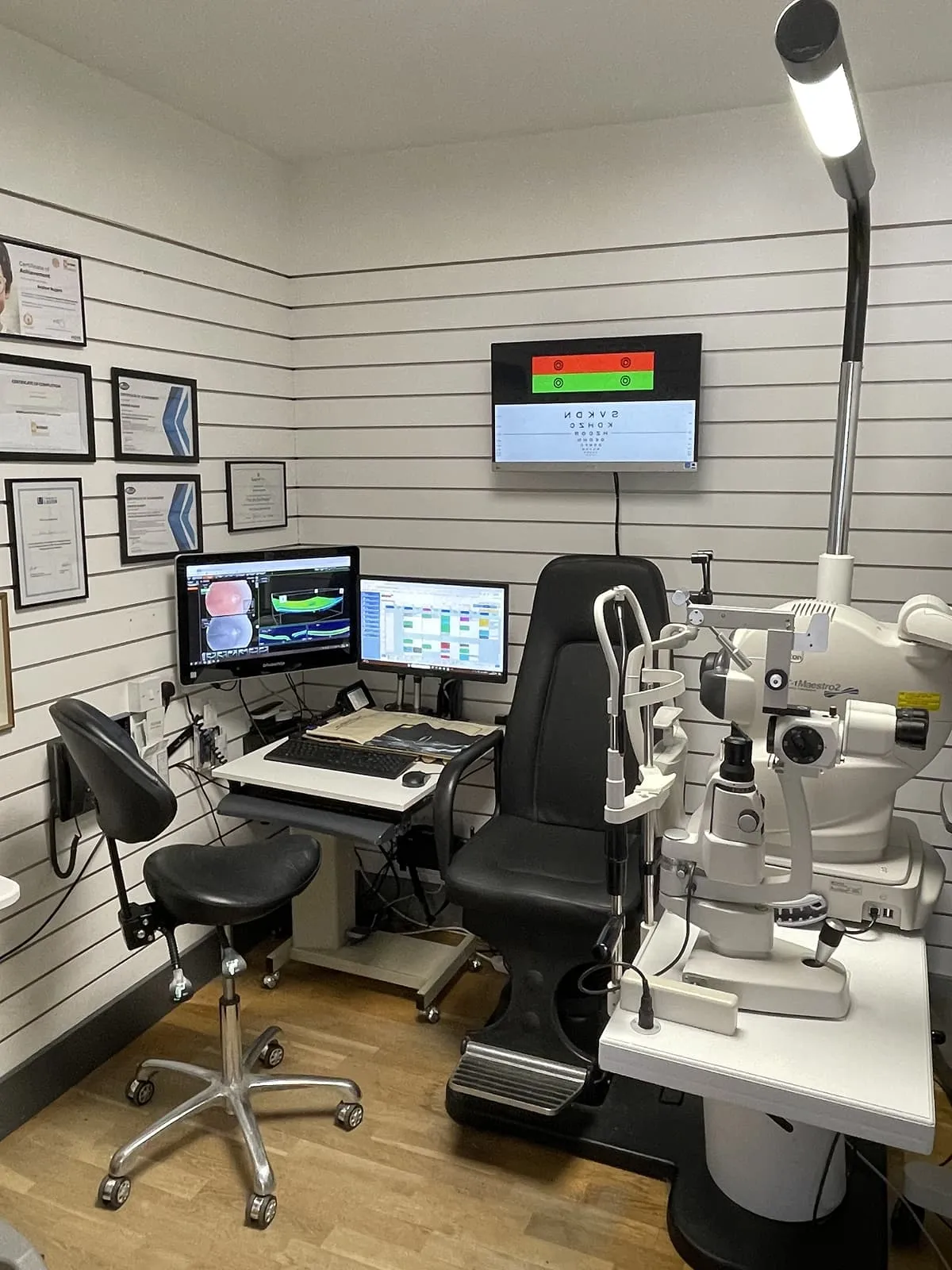 Interior of an optometry examination room with an eye testing chair, slit lamp, and two computer monitors displaying eye-related images and schedules.
