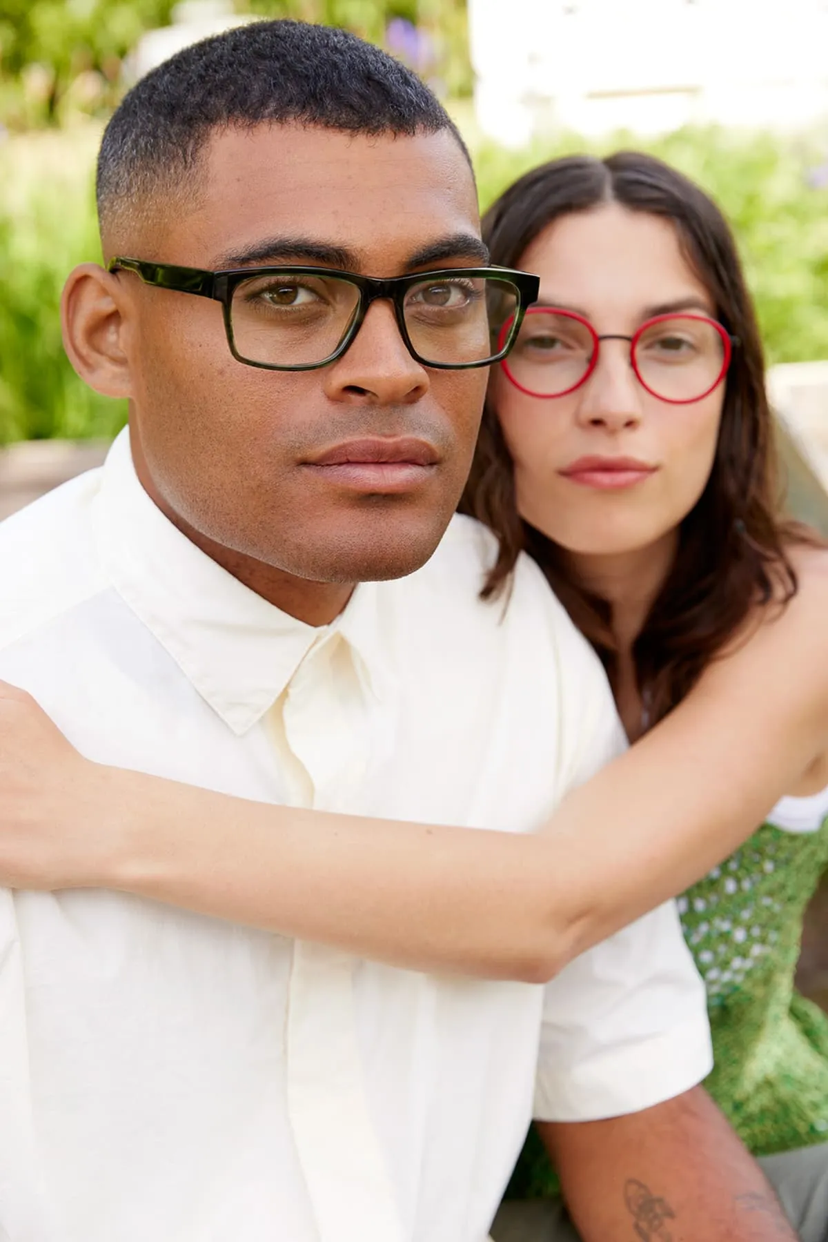 Close-up of a man wearing black rectangular glasses and a white shirt with a woman behind him wearing red round glasses and a green dress, her arm around his shoulder.