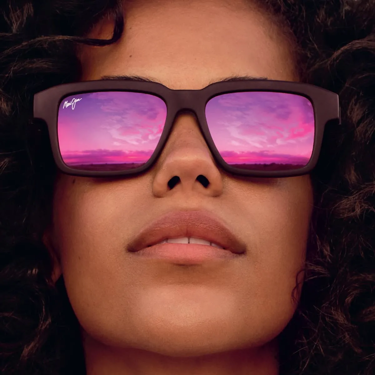 Close-up of a person wearing dark-framed sunglasses reflecting a pink and purple sunset sky.
