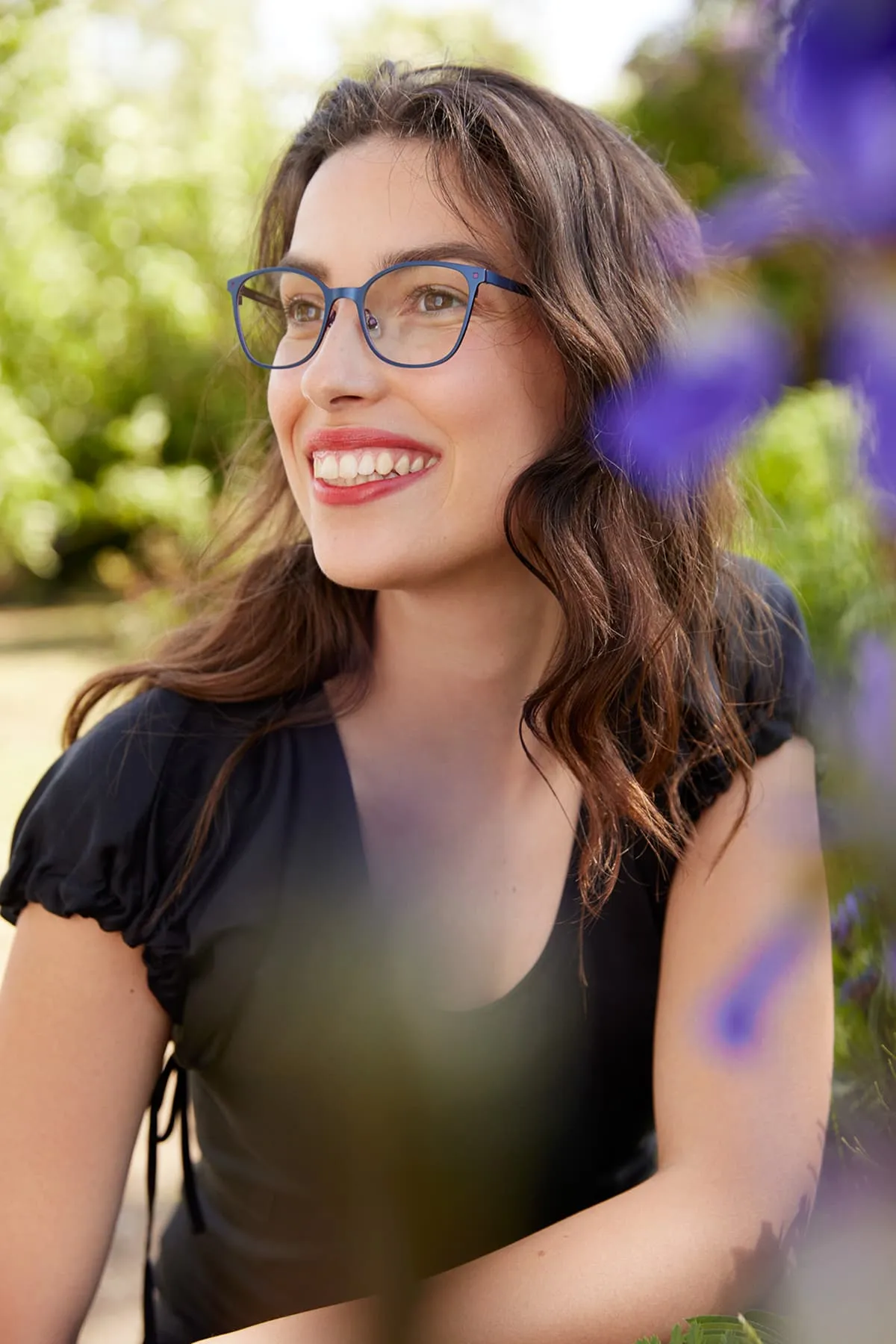 Smiling woman with wavy brown hair and blue glasses wearing a black top, outdoors with blurred greenery and purple flowers.