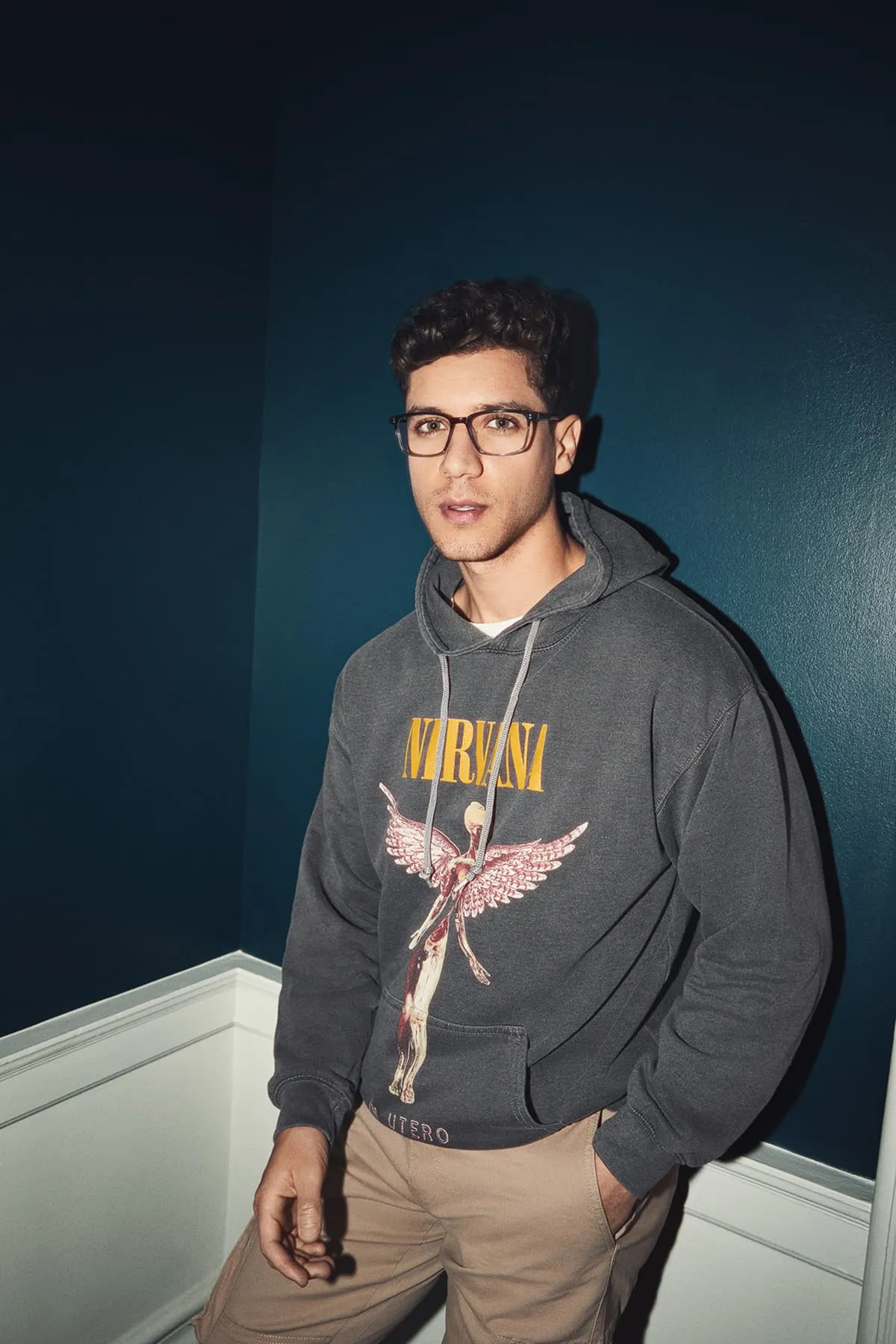 Young man with glasses wearing a dark gray Nirvana hoodie and beige pants, standing against a dark blue wall.