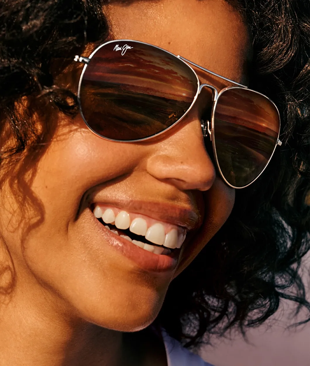 Close-up of a smiling woman wearing aviator sunglasses reflecting a sunset landscape.
