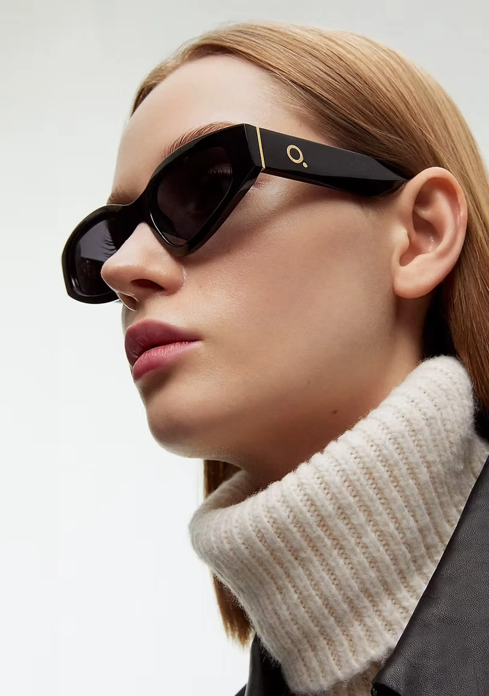 Close-up side profile of a woman wearing large black geometric sunglasses and a white ribbed turtleneck sweater.