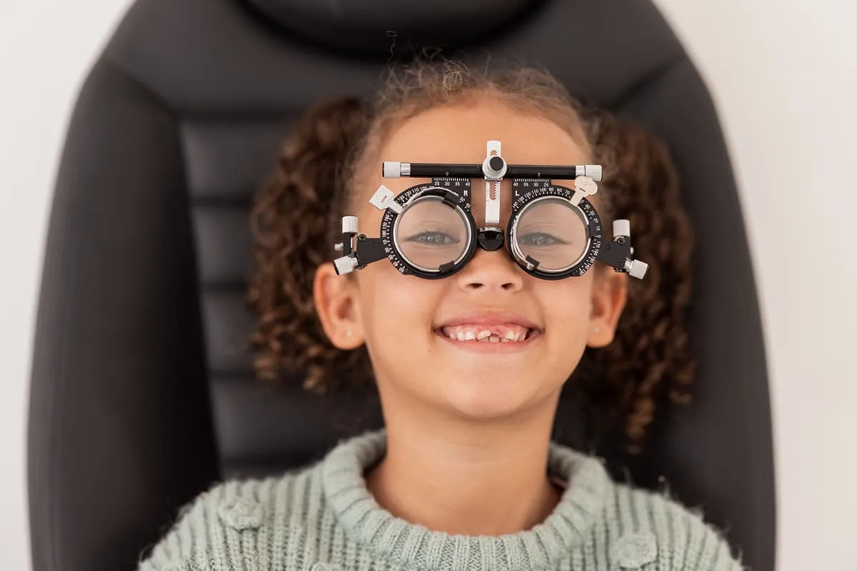 Smiling young girl wearing a trial frame for an eye exam while sitting in an eye exam chair.