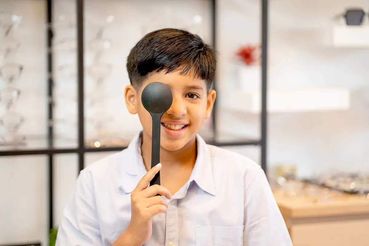 Smiling boy covering one eye with an eye occluder during an eye exam in an optical clinic.