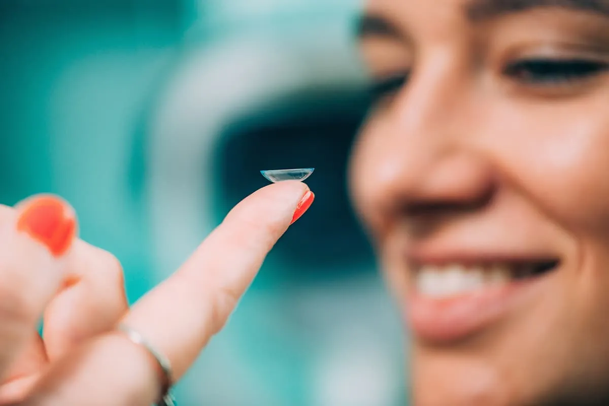 Close-up of a smiling woman holding a contact lens on her fingertip.