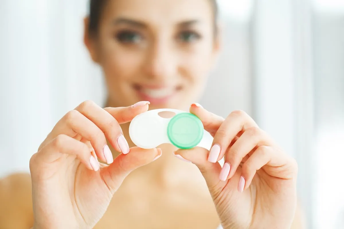 Smiling woman holding a white contact lens case with a green lid in front of her.