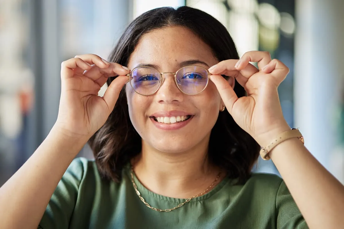 Smiling woman adjusting her round, clear glasses with blue light reflection, wearing a green top and necklace.