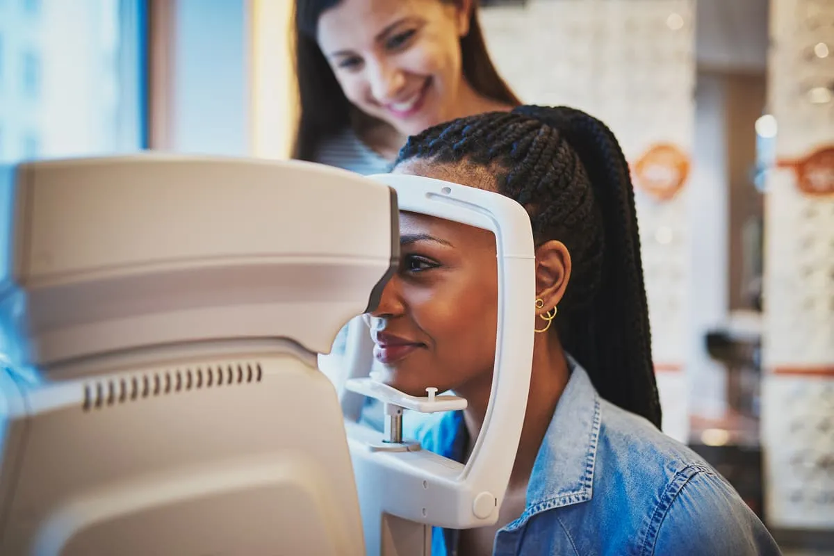 Woman undergoing an eye exam using an optical machine while another woman watches and smiles.
