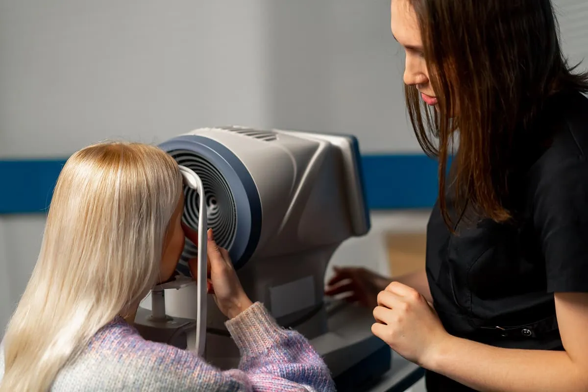 An eye care professional conducts an eye exam on a blonde woman using a specialized diagnostic machine.