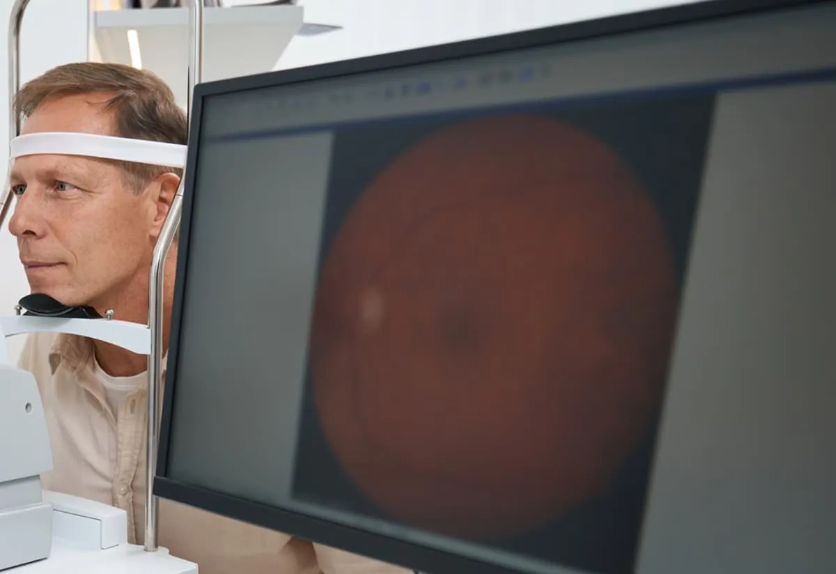 Man undergoing an eye examination using a machine with retina image displayed on a nearby monitor.