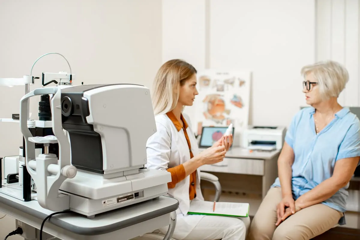 Eye examination room with an optical machine in the foreground and an eye doctor discussing with a female patient in the background.