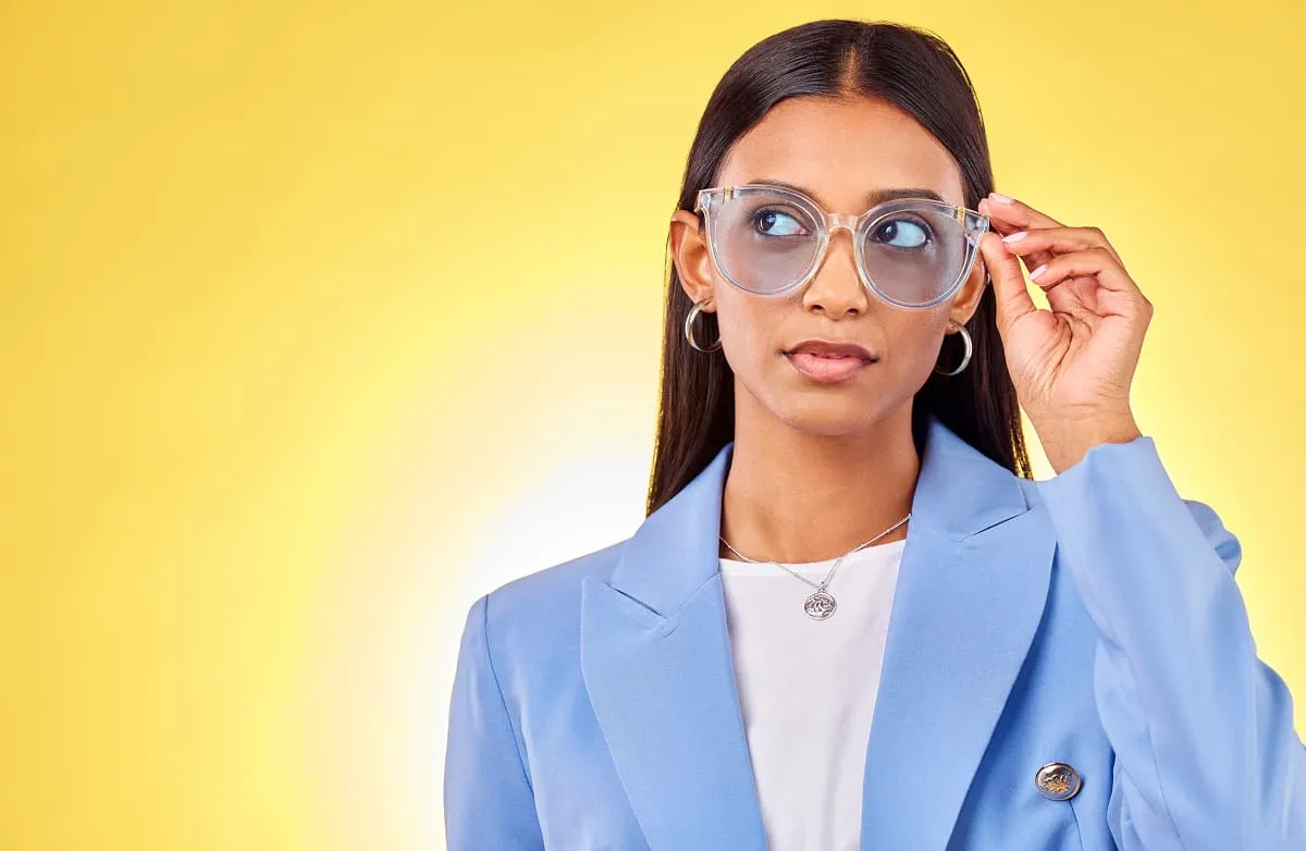 Woman wearing clear oversized glasses and a light blue blazer against a yellow background, adjusting her glasses while looking to the side.