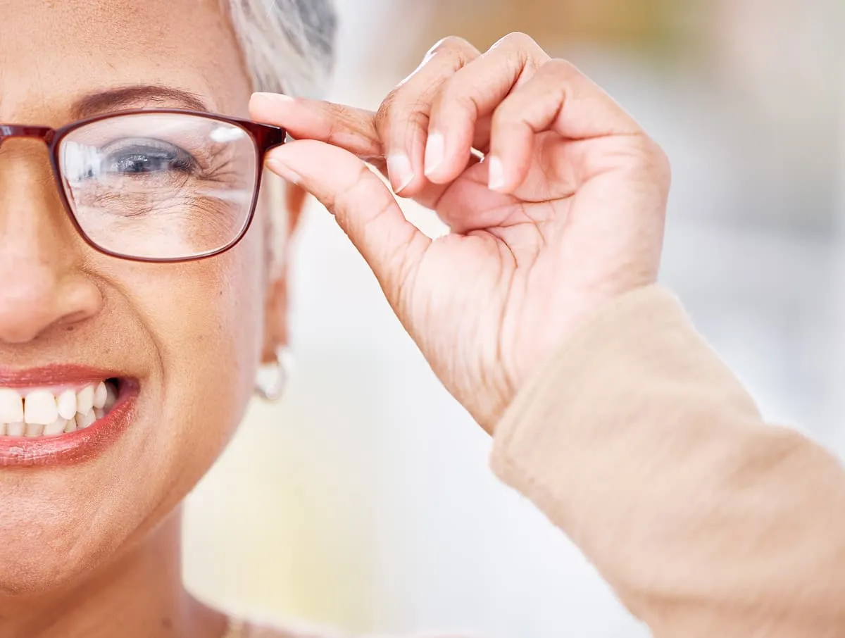 Close-up of a smiling older woman adjusting her brown-framed glasses with her right hand.