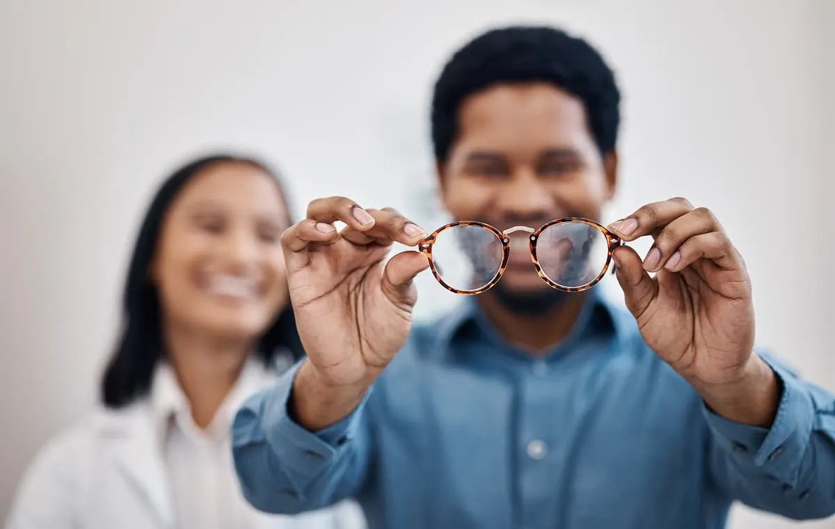 Man holding tortoiseshell eyeglasses in front of a smiling woman in a white coat.