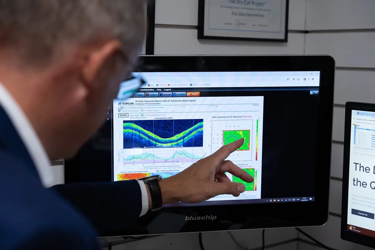 Optician pointing at an OCT retinal scan on a monitor, analysing diagnostic results for a patient eye examination.