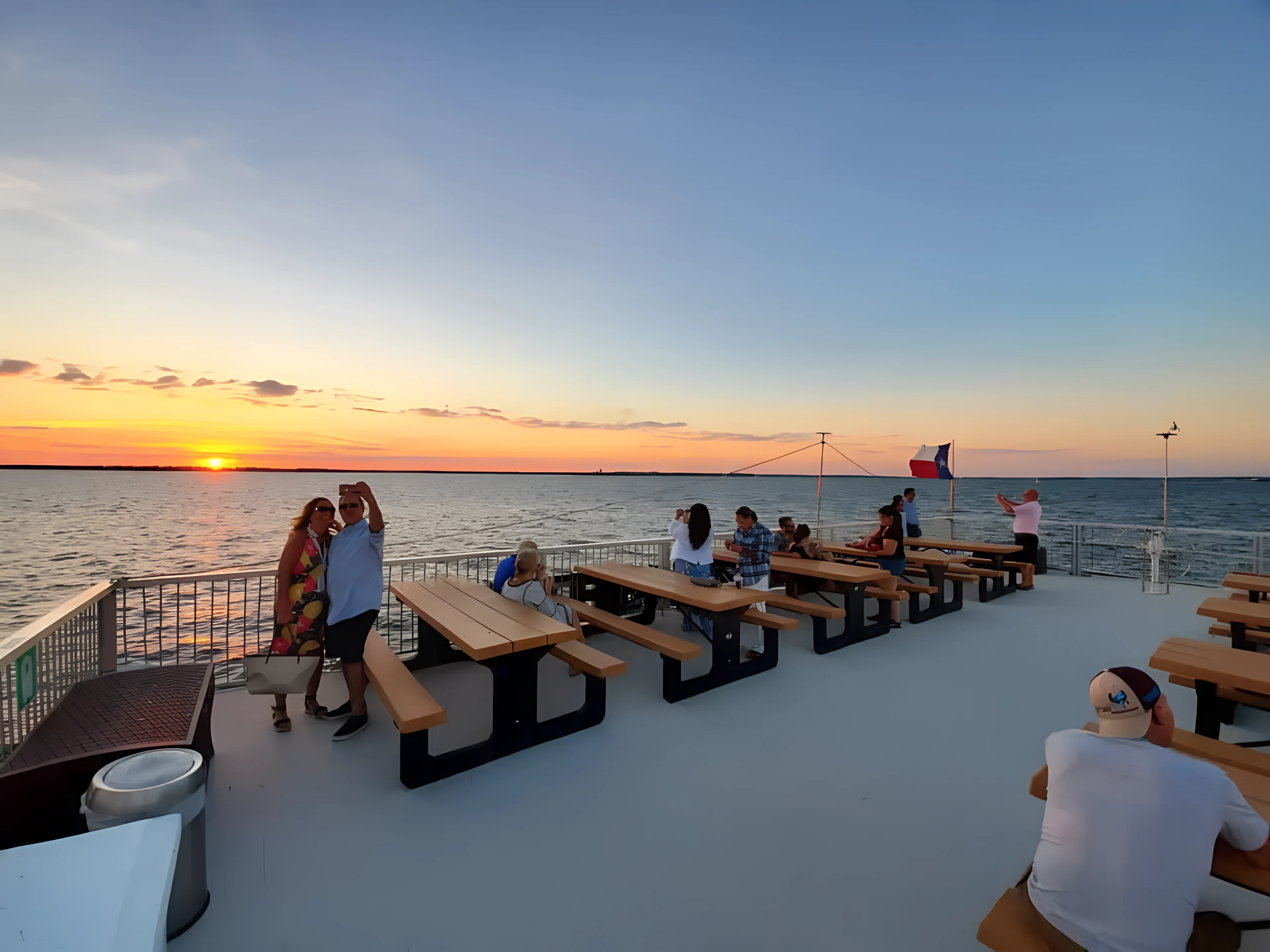 People sitting and taking photos on wooden benches on a boat deck at sunset over a calm body of water.