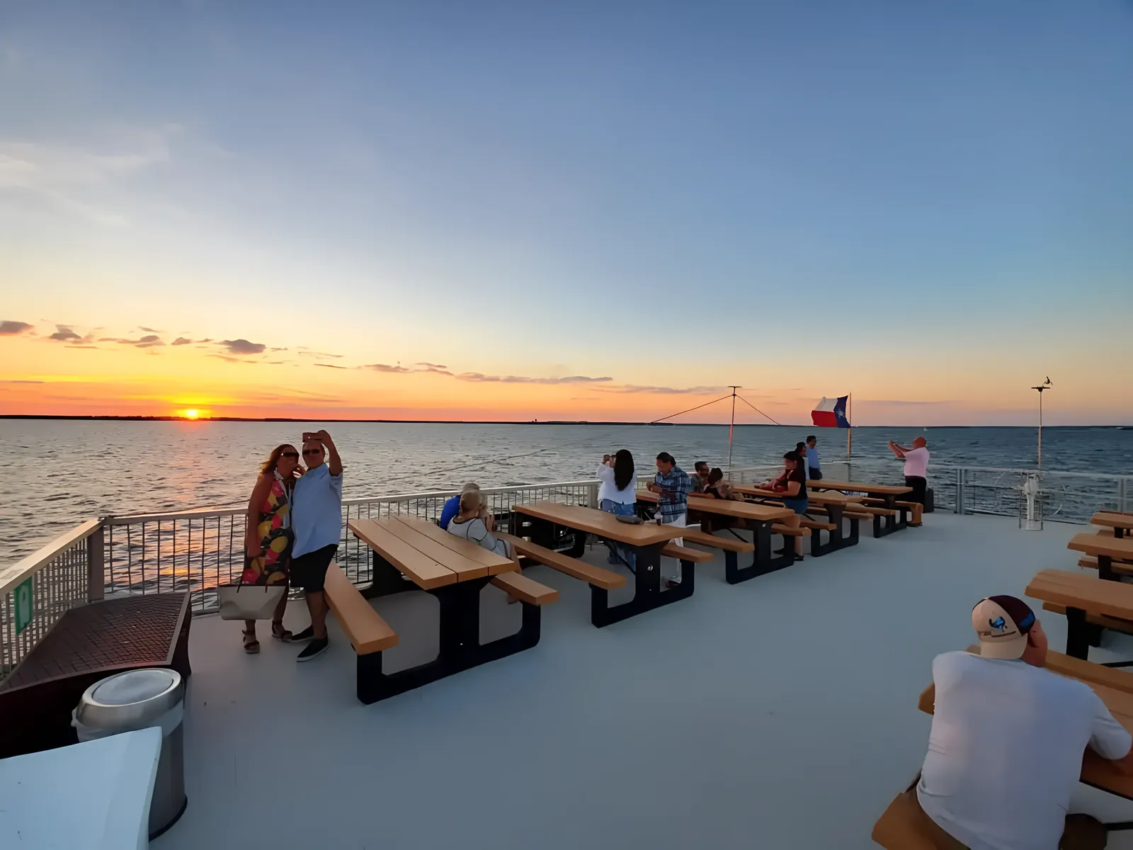 People sitting and taking photos on wooden benches on a boat deck at sunset over a calm body of water.