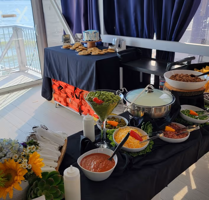 Buffet table with assorted food including salsa, shredded cheese, guacamole in a glass, seasoned meat, and chips, with a stack of cookies and a cake in the background near a sunlit window.