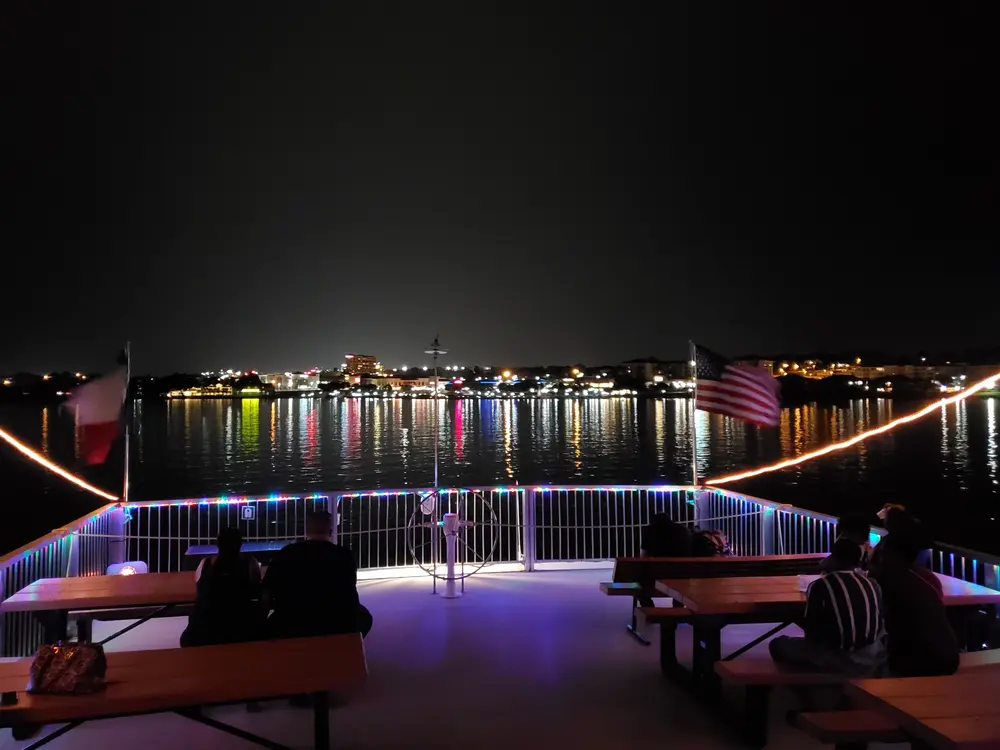 Buffet table with assorted food including salsa, shredded cheese, guacamole in a glass, seasoned meat, and chips, with a stack of cookies and a cake in the background near a sunlit window.