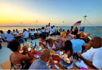 Group of people dining and socializing on a boat at sunset with American and Dominican flags.