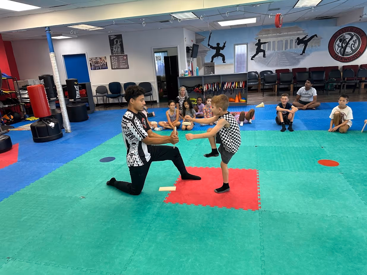Martial arts instructor holding a board for a young boy who is practicing a front kick in a training gym with other children seated in the background.