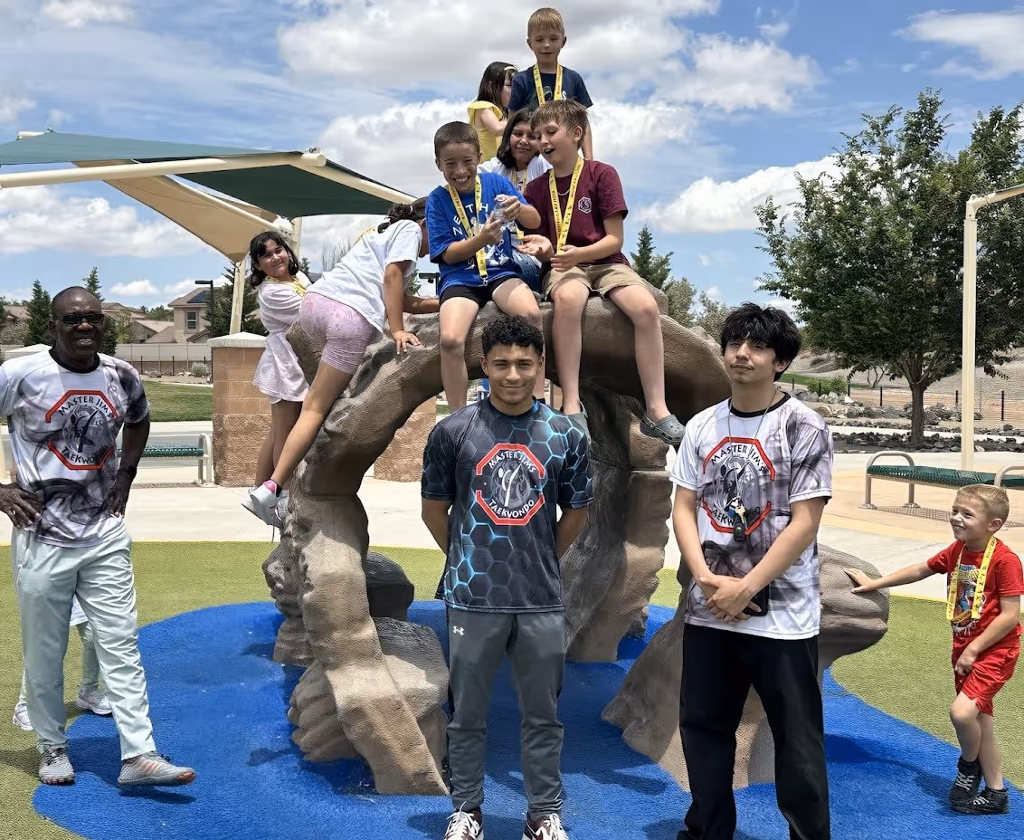 Group of children and three adults wearing Master Jim's Taekwondo shirts posing on and around a playground rock structure outdoors under a partly cloudy sky.