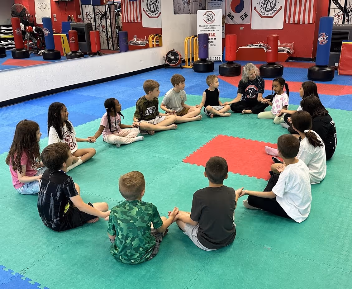 Group of children and an instructor sitting in a circle holding hands on a martial arts mat in a training studio.