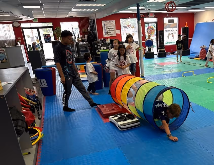 Children play in a martial arts gym with colorful mats, a tunnel toy, and training equipment while an instructor watches.