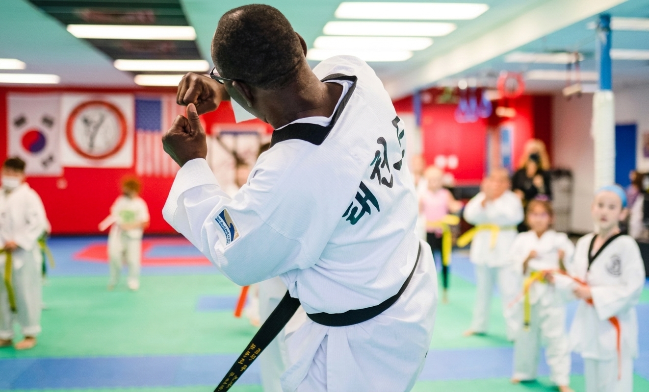 A high-angle shot from behind a martial arts instructor in a white dobok with a black belt, demonstrating a defensive stance in a brightly lit dojang while students in the background practice.