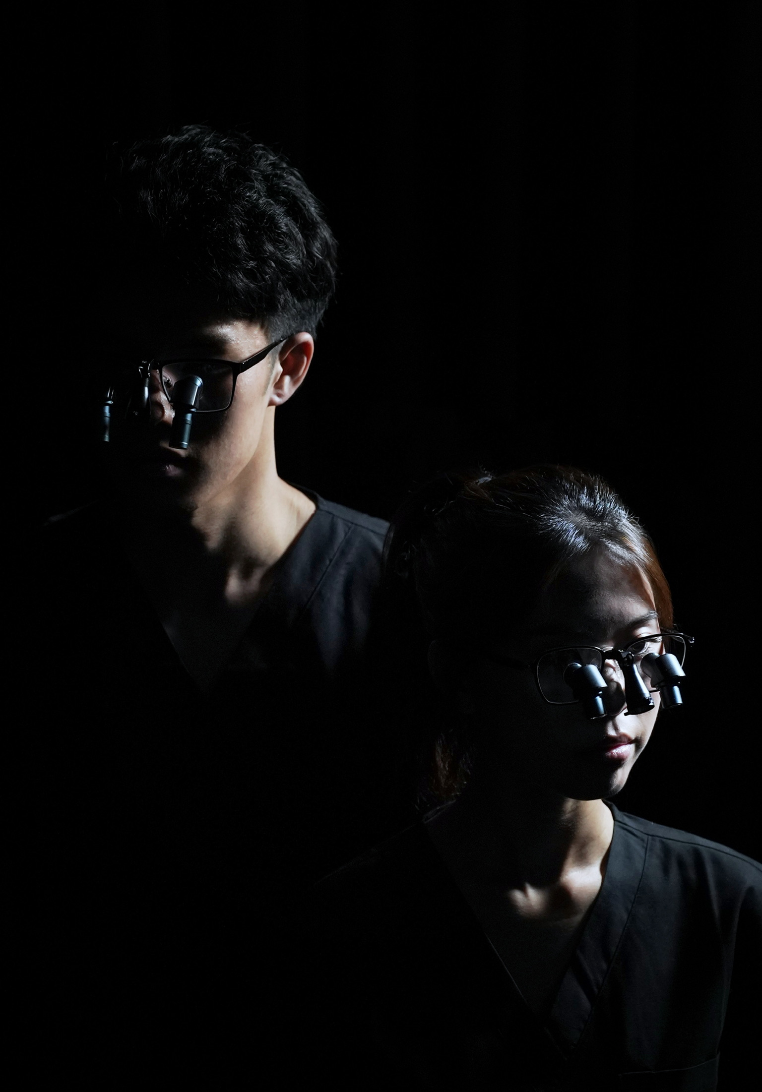 Two medical professionals in dark scrubs wearing prismara ergonomic loupes on their eyewear in low light.