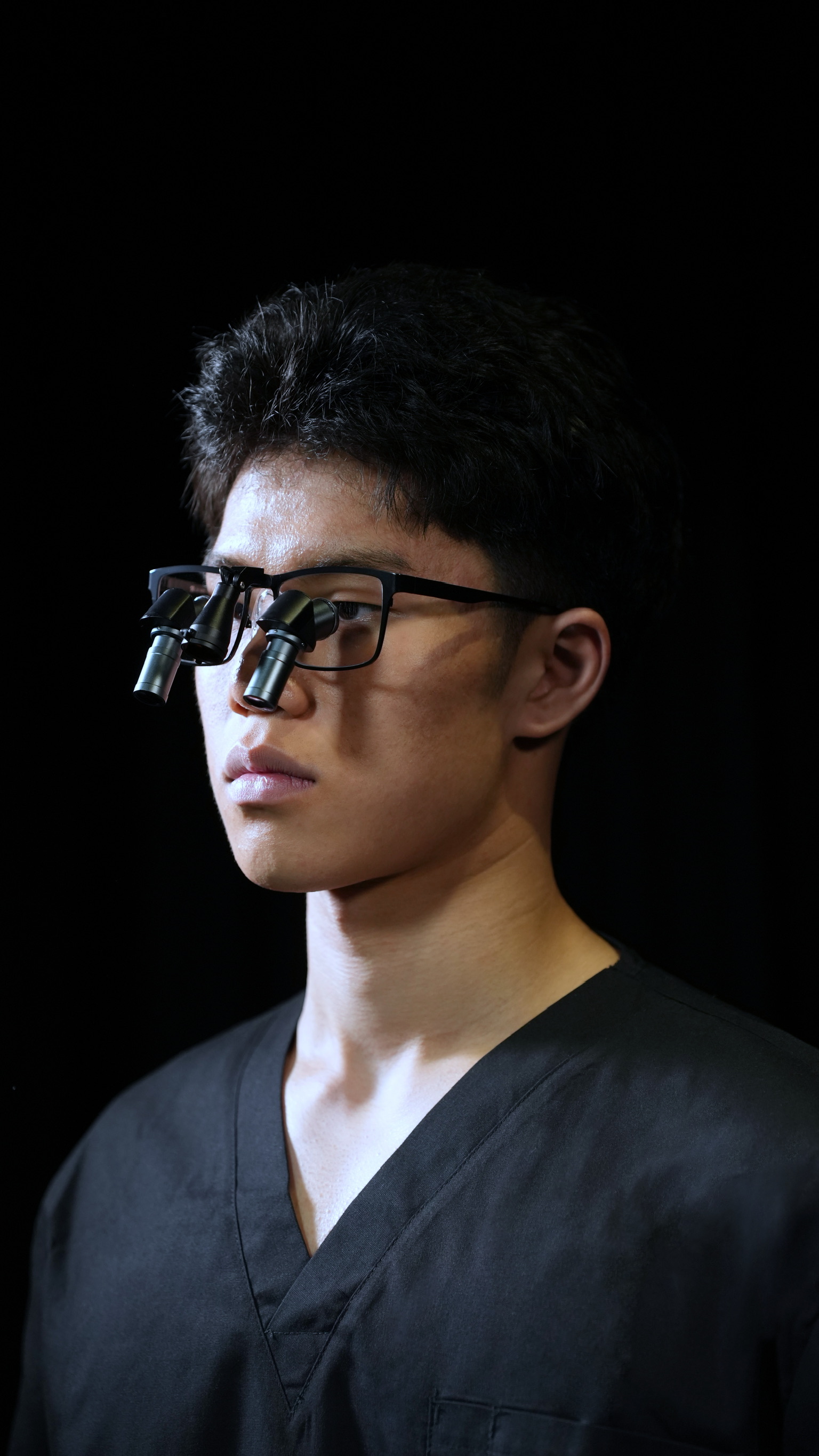 Young man wearing black medical scrubs and prismara ergonomic loupes with binocular lenses on a black background.