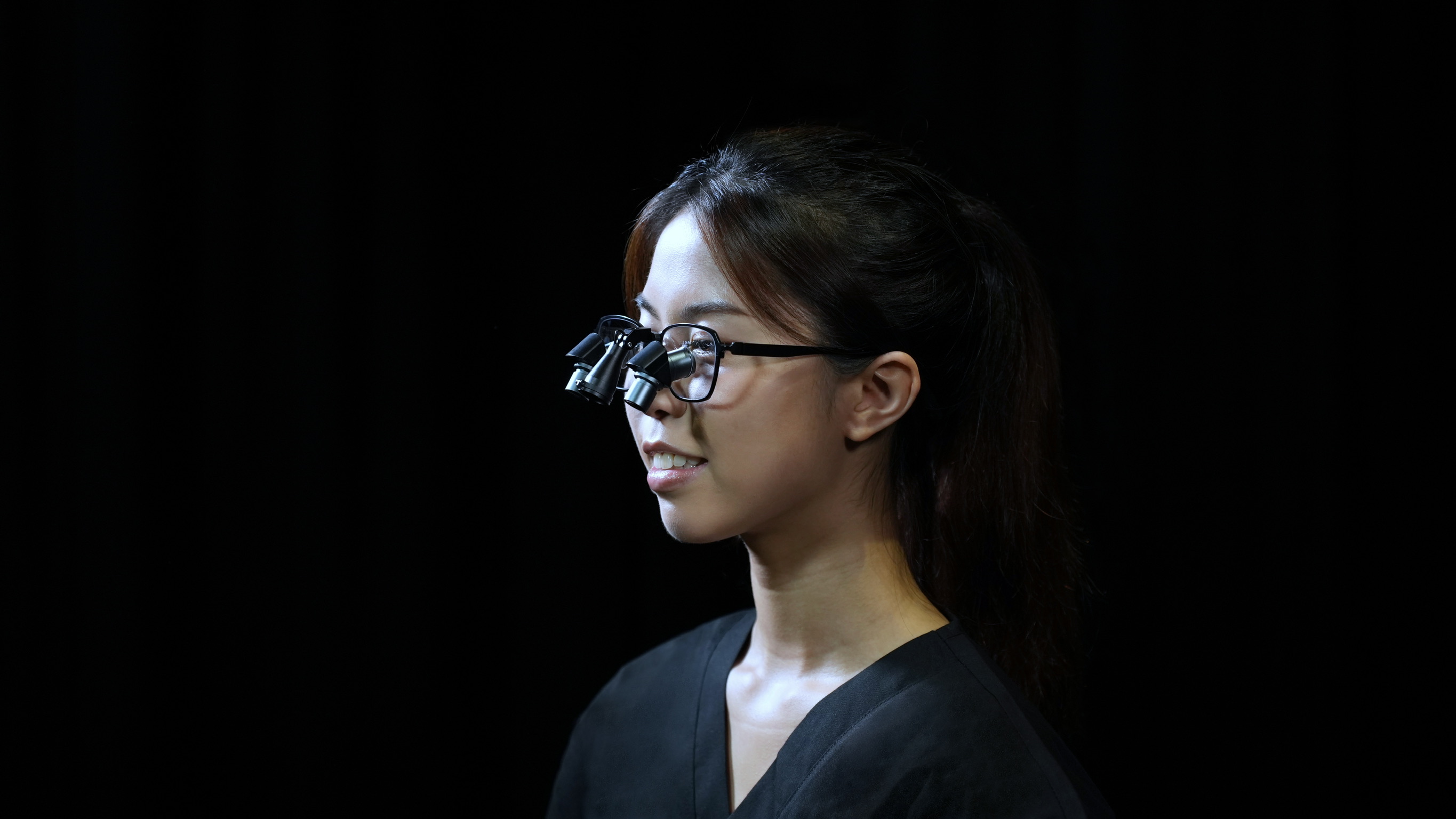 Side profile of a woman with glasses wearing Prismara ergonomic loupes against a black background.
