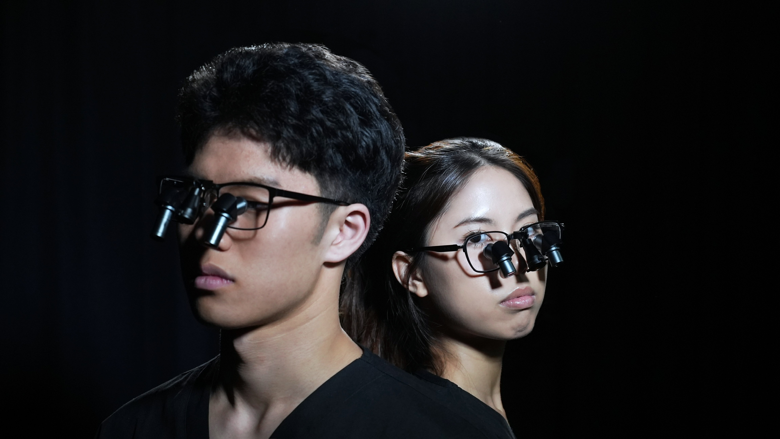 Man and woman wearing black prismara ergonomic loupes with headlamps against a dark background.