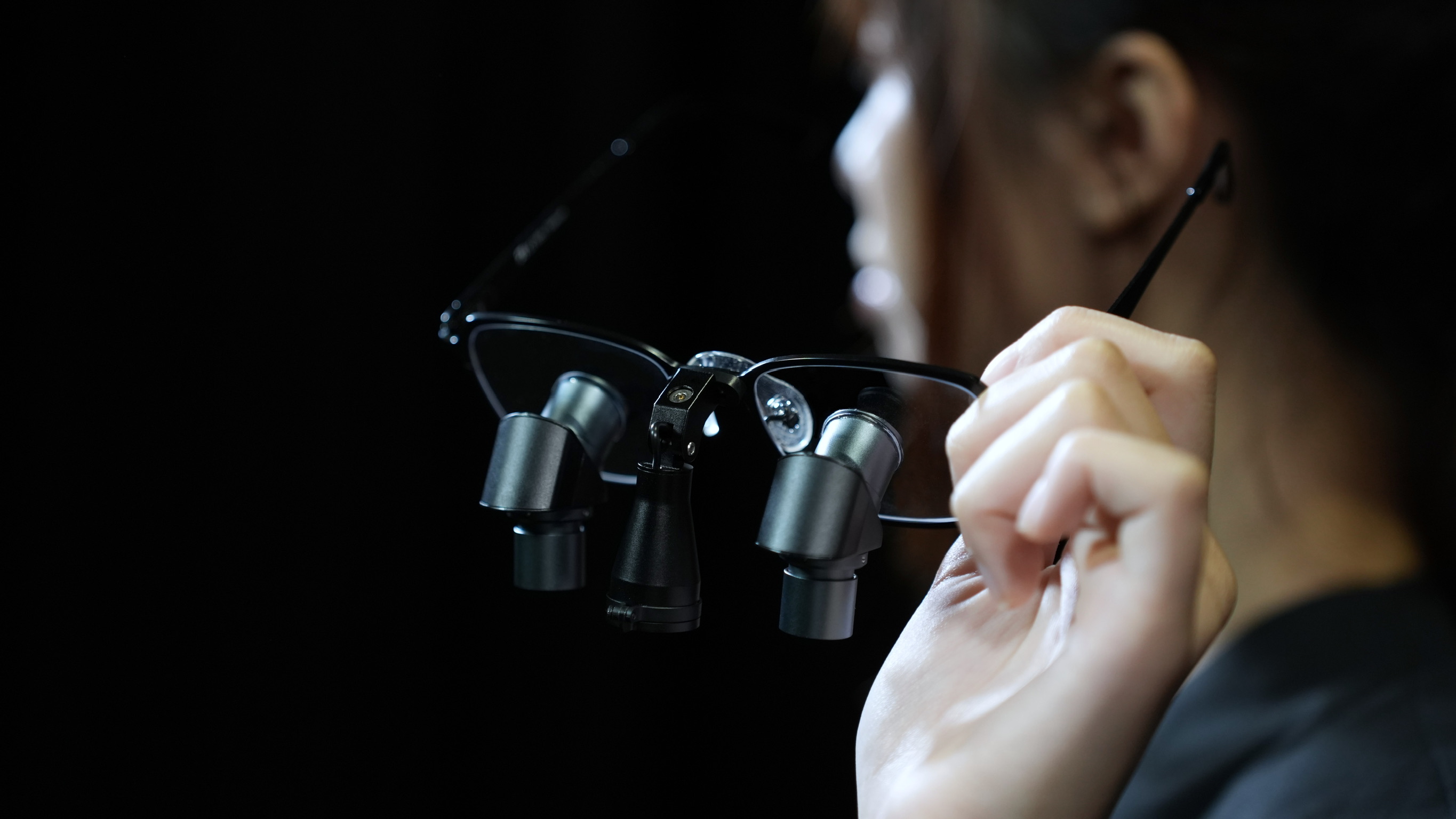 Close-up of a hand holding black prismara ergonomic loupes with dual lenses on the frames against a black background.