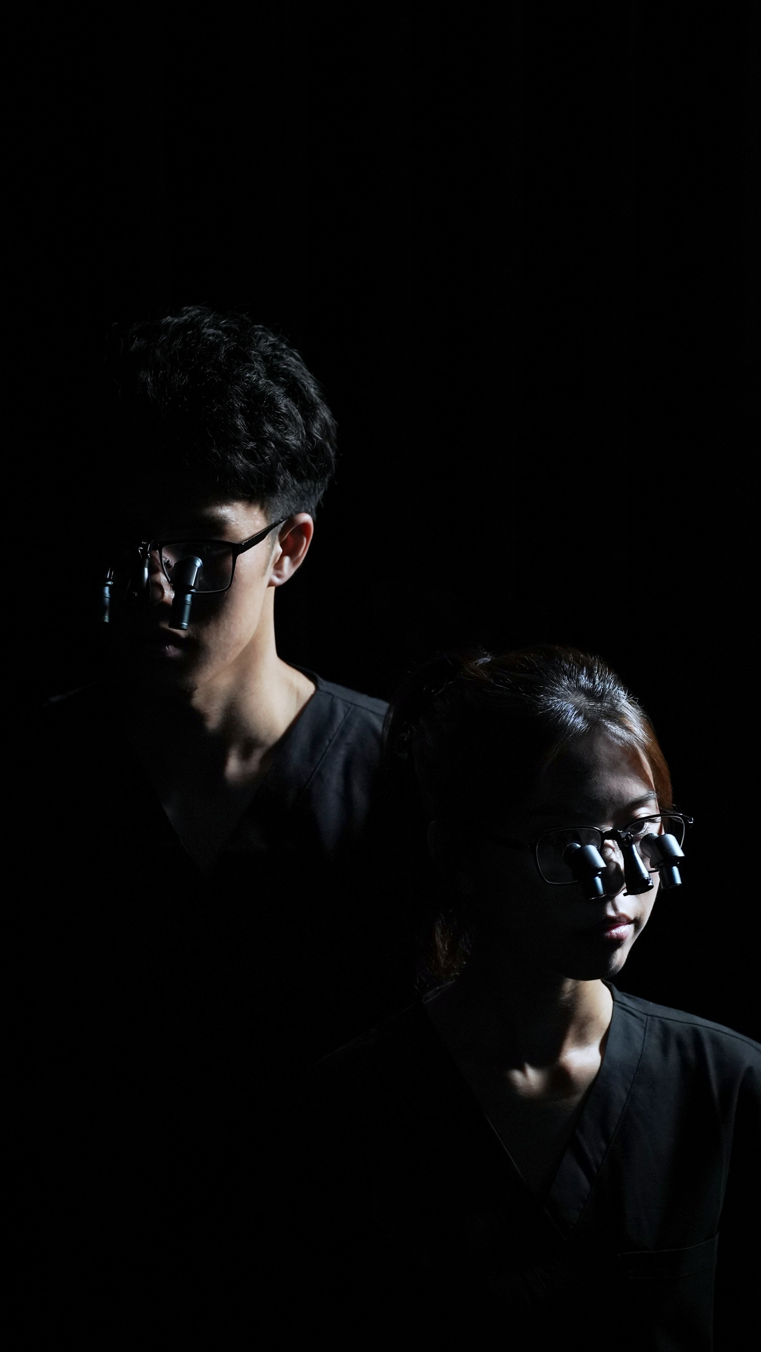 Two medical professionals wearing dark scrubs and prismara ergonomic loupes, partially illuminated against a dark background.