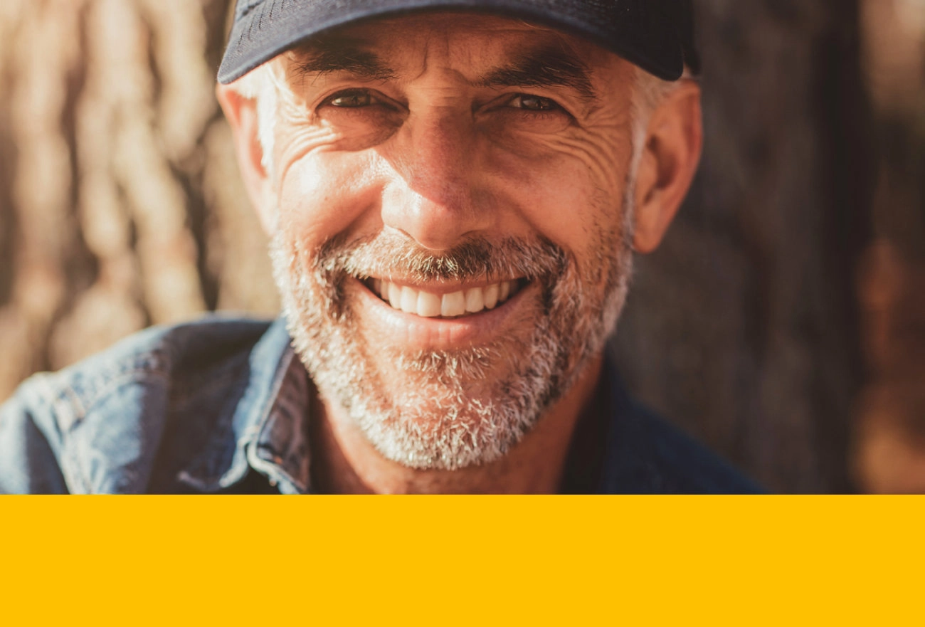 Older man with white beard wearing gray baseball cap smiling at camera with yellow band at bottom