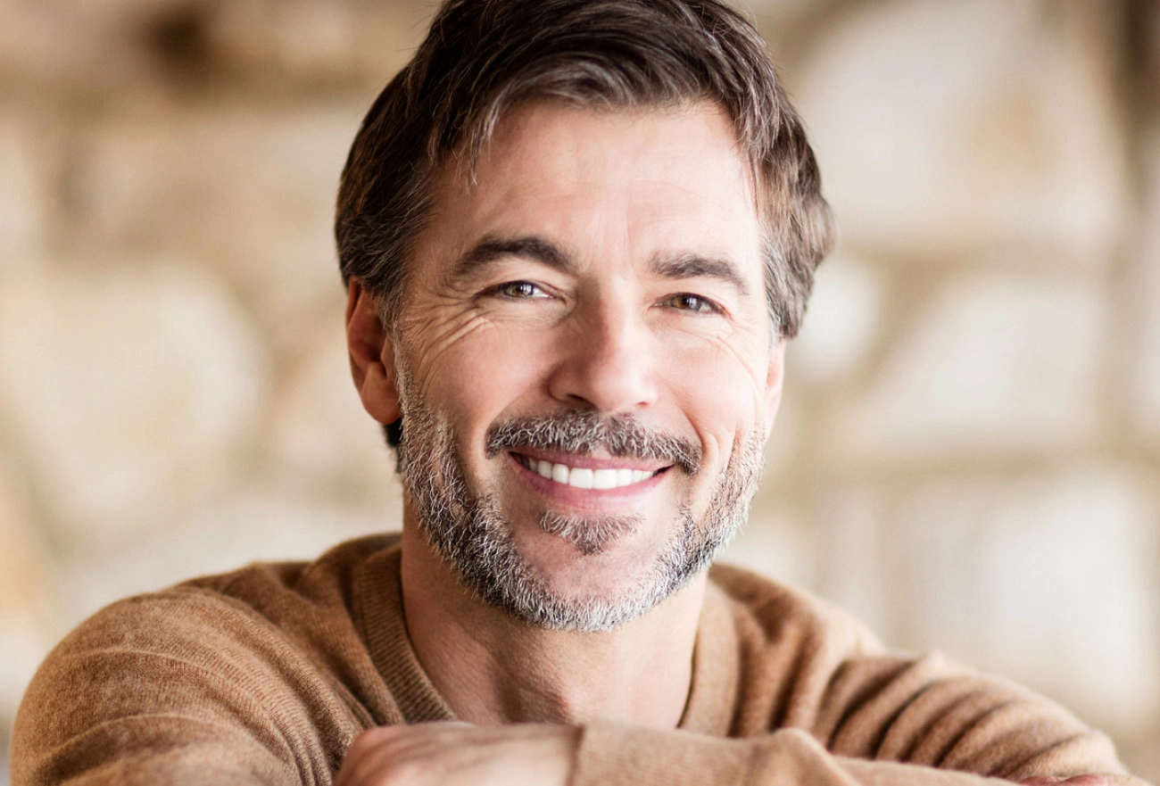 Man with gray beard wearing tan sweater smiling at camera against blurred background