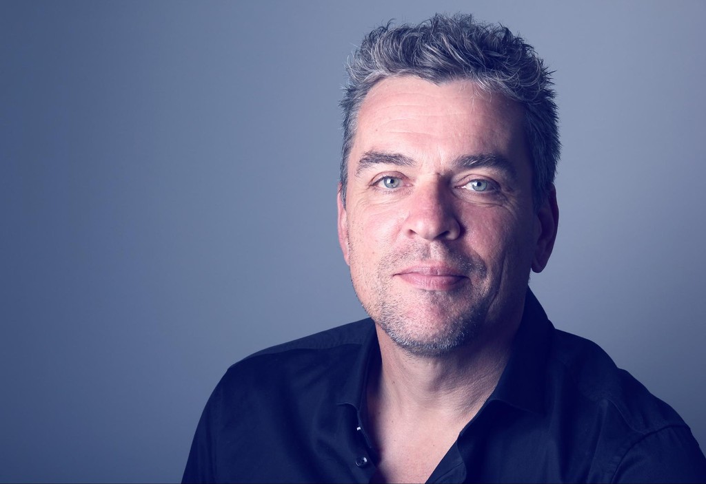 Man with gray and dark hair wearing dark blue shirt against blue background looking at camera