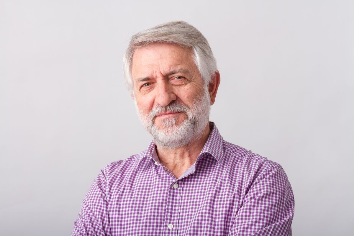 Older man with white beard wearing purple checkered shirt against neutral background
