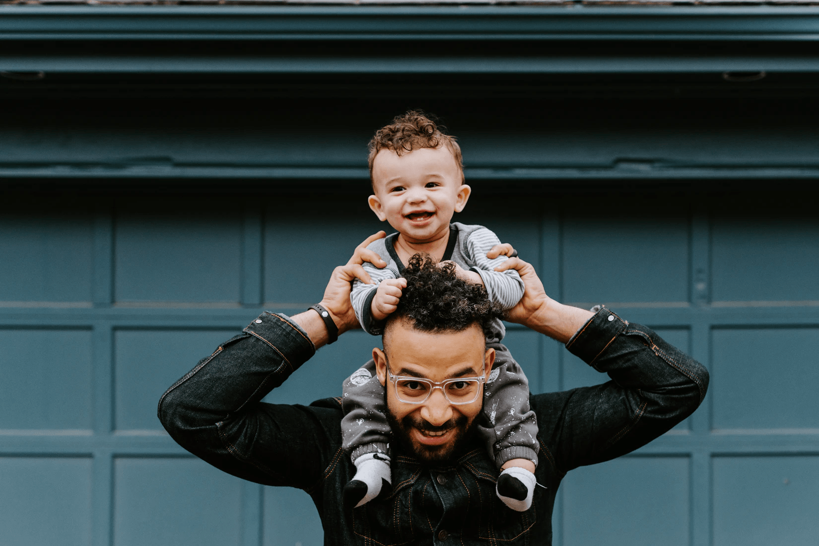 Man wearing glasses holding smiling toddler on shoulders in front of blue garage door