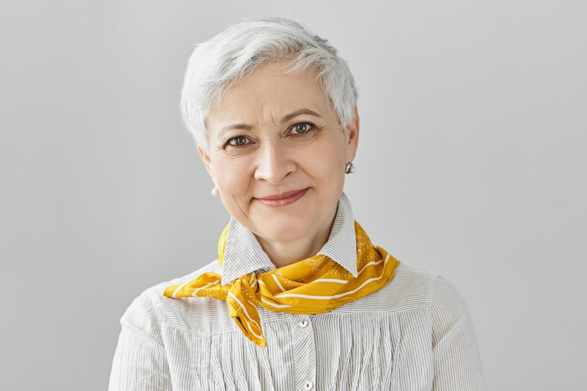 Older woman with short white hair wearing white striped shirt and yellow scarf smiling at camera