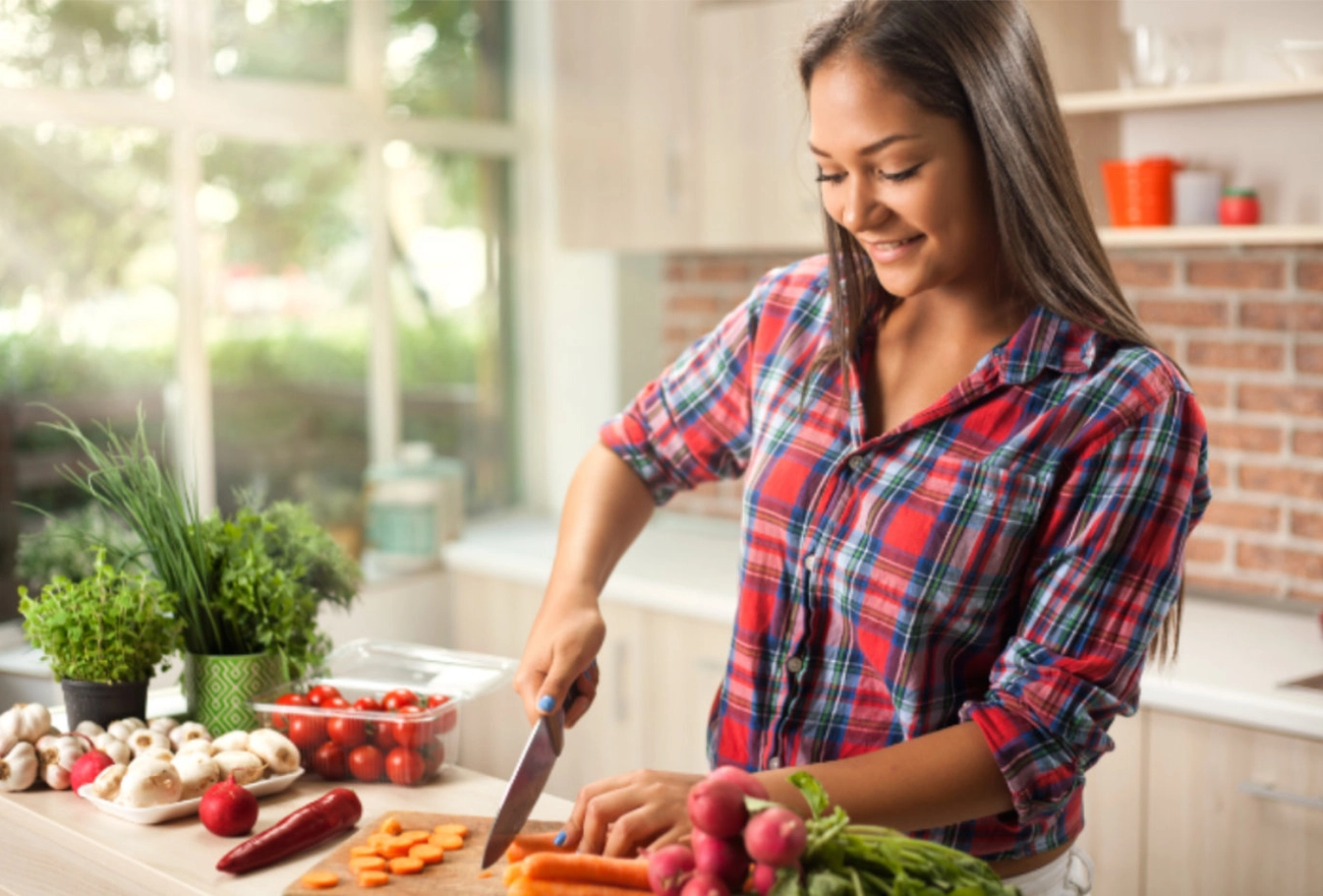 Woman in plaid shirt chopping fresh vegetables in bright kitchen with herbs and produce